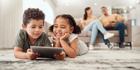 un bambino e una bambina giocano con il tablet con i genitori sullo sfondo