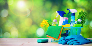 Cestino con detergenti spray, panni in microfibra, spugna e guanti per pulizie domestiche su tavolo, sfondo verde luminoso.