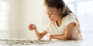 Bambina concentrata mentre osserva e sceglie un tassello tra i pezzi di un puzzle disposti su un tavolo chiaro.