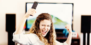 Ragazza sorridente alza un telecomando davanti a una TV accesa, esprimendo entusiasmo in un ambiente domestico.