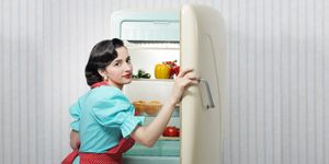 Donna con abito in stile vintage apre un frigorifero retrò con verdure fresche e alimenti ordinati all’interno.