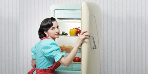 Donna con abito in stile vintage apre un frigorifero retrò con verdure fresche e alimenti ordinati all’interno.