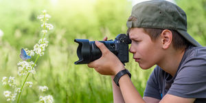 Ragazzo con cappellino fotografa una farfalla su un fiore in un prato verde, utilizzando una fotocamera con teleobiettivo.