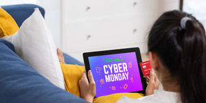 Persona sul divano usa un tablet con schermata Cyber Monday mentre tiene una carta di pagamento per acquistare online