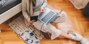 Persona seduta a terra accanto al letto che usa un laptop collegato a una stampante portatile con foto sparse sul tappeto.