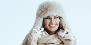 Donna sorridente con colbacco e guanti in lana tiene in mano un barattolo di crema viso su sfondo chiaro invernale.