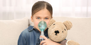 Bambina seduta sul divano usa un aerosol tenendo in braccio un orsetto di peluche.