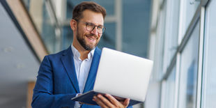 Uomo sorridente in giacca blu osserva lo schermo del suo laptop in un ambiente luminoso con grandi vetrate.
