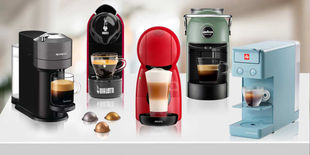 Cinque macchine da caffè colorate in fila con tazzine pronte e capsule assortite sparse sul piano bianco.