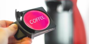 Capsula di caffè tenuta in mano davanti a macchina per macchine da caffè in capsule, con scritta coffee visibile