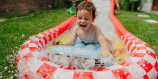 Bambino sorridente scivola sull’acqua in giardino su una corsia gonfiabile con bordi rossi e bianchi.