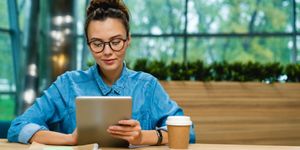 Donna con occhiali e camicia di jeans usa un tablet seduta a un tavolo, accanto a un bicchiere di caffè d’asporto.
