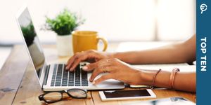 Una persona digita sulla tastiera di un laptop su un tavolo di legno con tazza gialla, occhiali e pianta accanto.