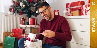 Uomo sorridente apre un regalo davanti all’albero di Natale, circondato da pacchi colorati e decorazioni festive.