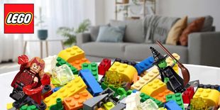Mattoncini colorati sparsi in primo piano con due minifigure in posa davanti a un logo su sfondo di salotto moderno.