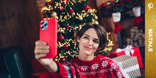 Ragazza con maglione rosso che scatta un selfie vicino all’albero decorato, tra regali colorati e atmosfera festiva.