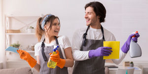 Coppia sorridente con guanti e grembiuli tiene detergenti e panni mentre si dedica alle pulizie domestiche in ambiente luminoso.