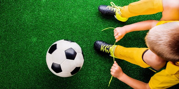 Un bambino in divisa sportiva allaccia le scarpe da calcio nere con lacci gialli, accanto a un pallone su prato verde.