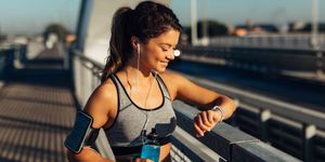 Donna in abbigliamento sportivo sorride mentre controlla l’orologio da polso, con auricolari e borraccia durante l’allenamento.