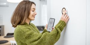 Donna regola un termostato smart a muro mostrando sullo smartphone la stessa temperatura impostata.