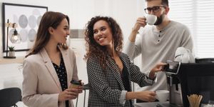 Tre colleghi sorridenti in ufficio preparano e bevono caffè insieme, riuniti attorno a una macchina automatica moderna.