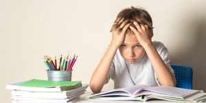Bambino seduto alla scrivania con le mani tra i capelli, davanti a quaderni aperti e matite colorate, con espressione stanca.