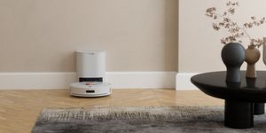 Robot lavapavimenti bianco in ricarica alla base, posizionato su pavimento in legno accanto a un tavolino moderno.