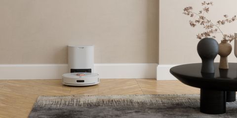 Robot lavapavimenti bianco in ricarica alla base, posizionato su pavimento in legno accanto a un tavolino moderno.