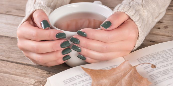 Mani con unghie smaltate verde scuro lucido stringono una tazza di tè caldo, accanto a libro aperto e foglia secca autunnale.