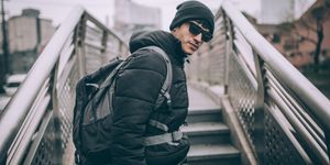 Uomo con giacca imbottita nera, berretto e zaino, su una scalinata urbana in giornata grigia.