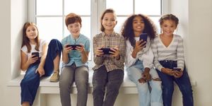 Cinque bambini seduti su un davanzale sorridono guardando gli schermi dei loro smartphone in un ambiente luminoso.