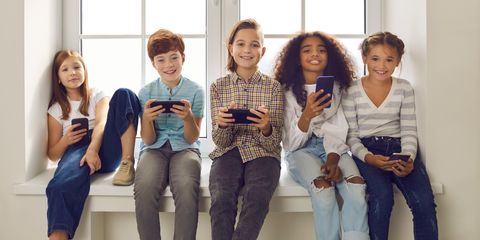 Cinque bambini seduti su un davanzale sorridono guardando gli schermi dei loro smartphone in un ambiente luminoso.