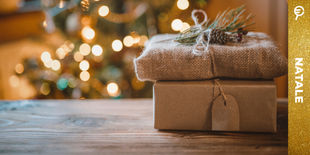 Pacchi regalo in carta e juta decorati con pigna e ramoscello su tavolo di legno, con luci natalizie sfocate sullo sfondo.