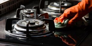 Mani con guanti arancioni passano un panno sui bruciatori dei fornelli a gas, rimuovendo sporco e incrostazioni.