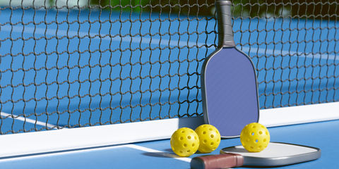 Racchetta da pickleball appoggiata alla rete con tre palline gialle forate su campo azzurro illuminato dal sole