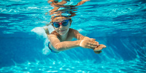 Bambino che nuota sott’acqua in piscina con occhialini, corpo disteso e braccia in avanti, illuminato dalla luce riflessa sull’acqua.