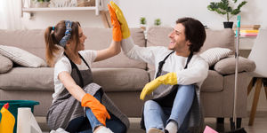 Coppia seduta sul pavimento festeggia le pulizie con il cinque, indossando guanti e grembiuli in salotto