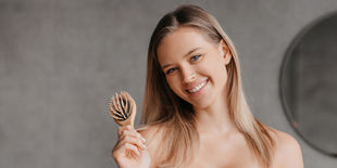 Donna sorridente con capelli lisci tiene in mano una spazzola di legno davanti a una parete grigia e uno specchio sfocato.