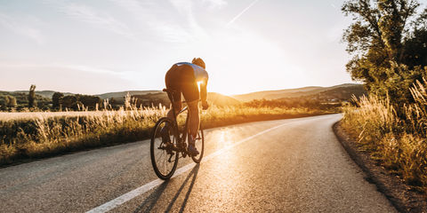 Ciclista pedala su strada panoramica al tramonto, silhouette illuminata dal sole con campi e colline sullo sfondo