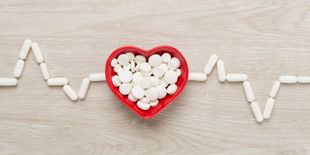 Ciotola rossa a forma di cuore piena di compresse bianche, circondata da capsule disposte come tracciato cardiaco.