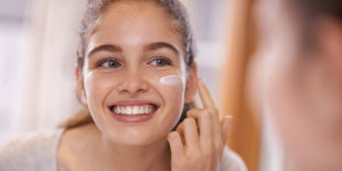 Ragazza sorridente davanti allo specchio applica una crema sulla guancia con le dita, in bagno luminoso durante la routine viso quotidiana.