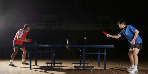 Due giocatori impegnati in una partita su tavolo da ping pong in palestra, illuminati da luce soffusa e concentrati sullo scambio