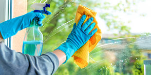 Mani con guanti blu puliscono una finestra con panno giallo e detergente spray, lasciando il vetro brillante con sfondo verde sfocato