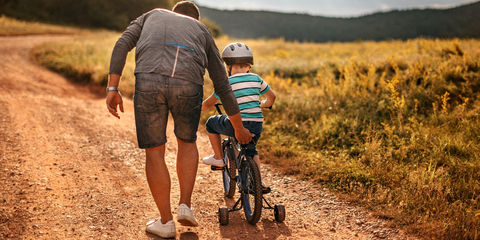 Adulto accompagna un bambino in bici con rotelle su un sentiero sterrato al tramonto, tra prati e colline sullo sfondo