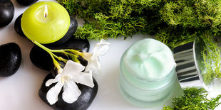 Vasetto di crema cosmetica tra pietre nere, fiori bianchi, muschio verde e candela accesa